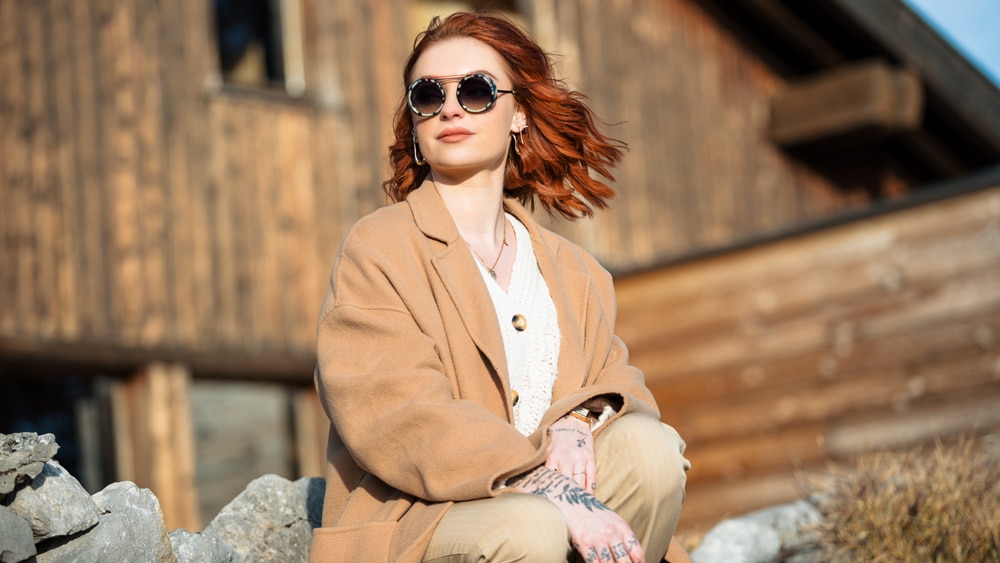 Young woman wearing La Petite Lunette Rouge sunglasses, outside, in front of a mountain chalet